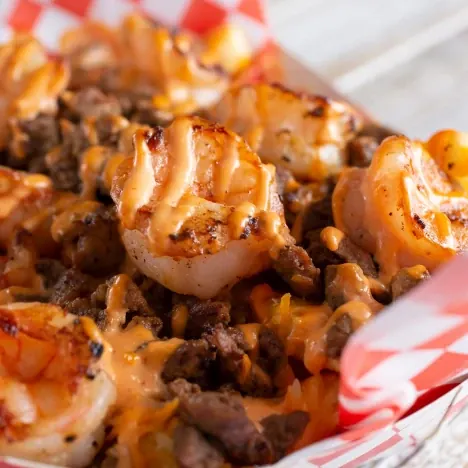 A basket of fries with steak and shrimp on top layered with a drizzle of cheese
